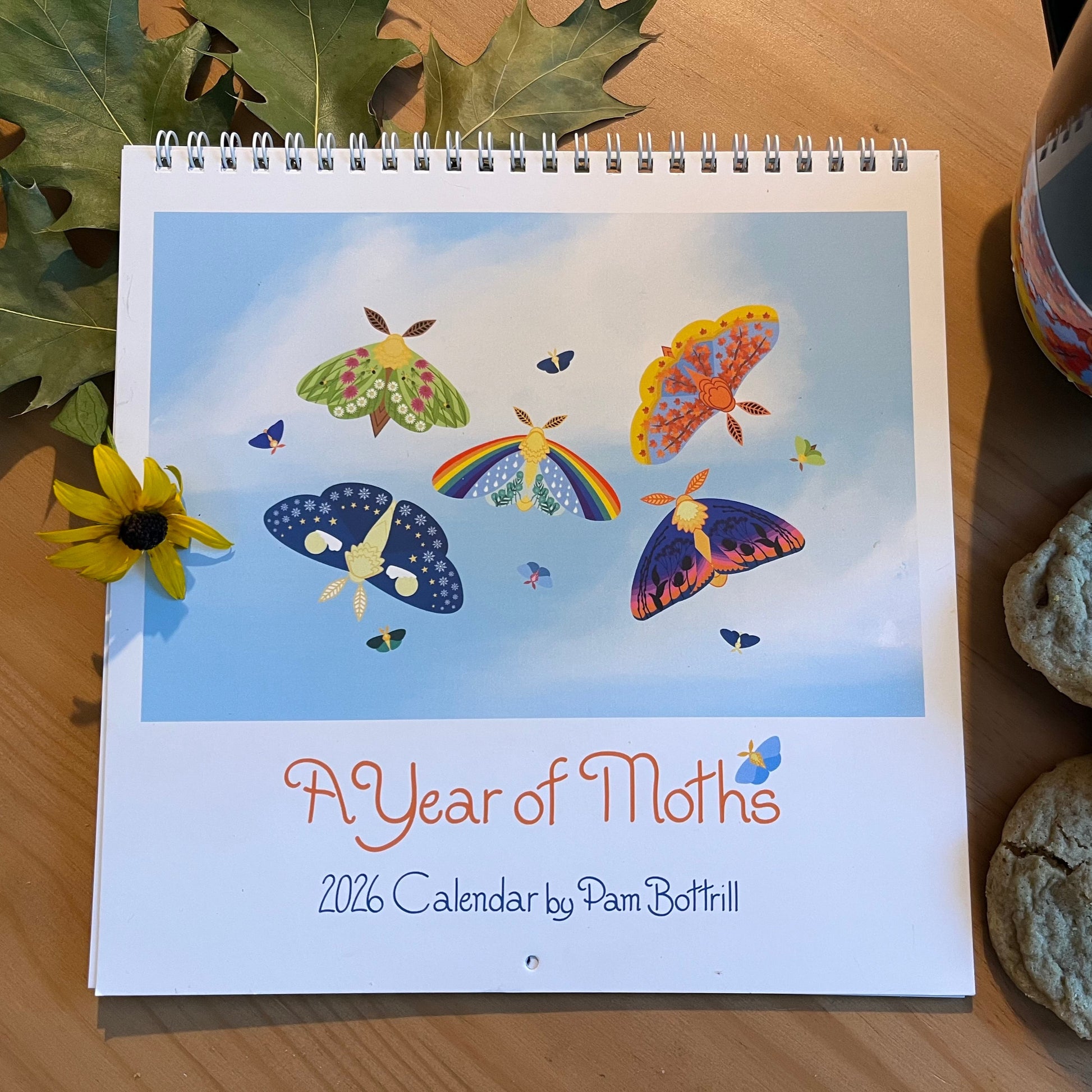 A Year of Moths Calendar rests on wooden table surrounded by a coffee mug, cookies, leaves and a flower 
