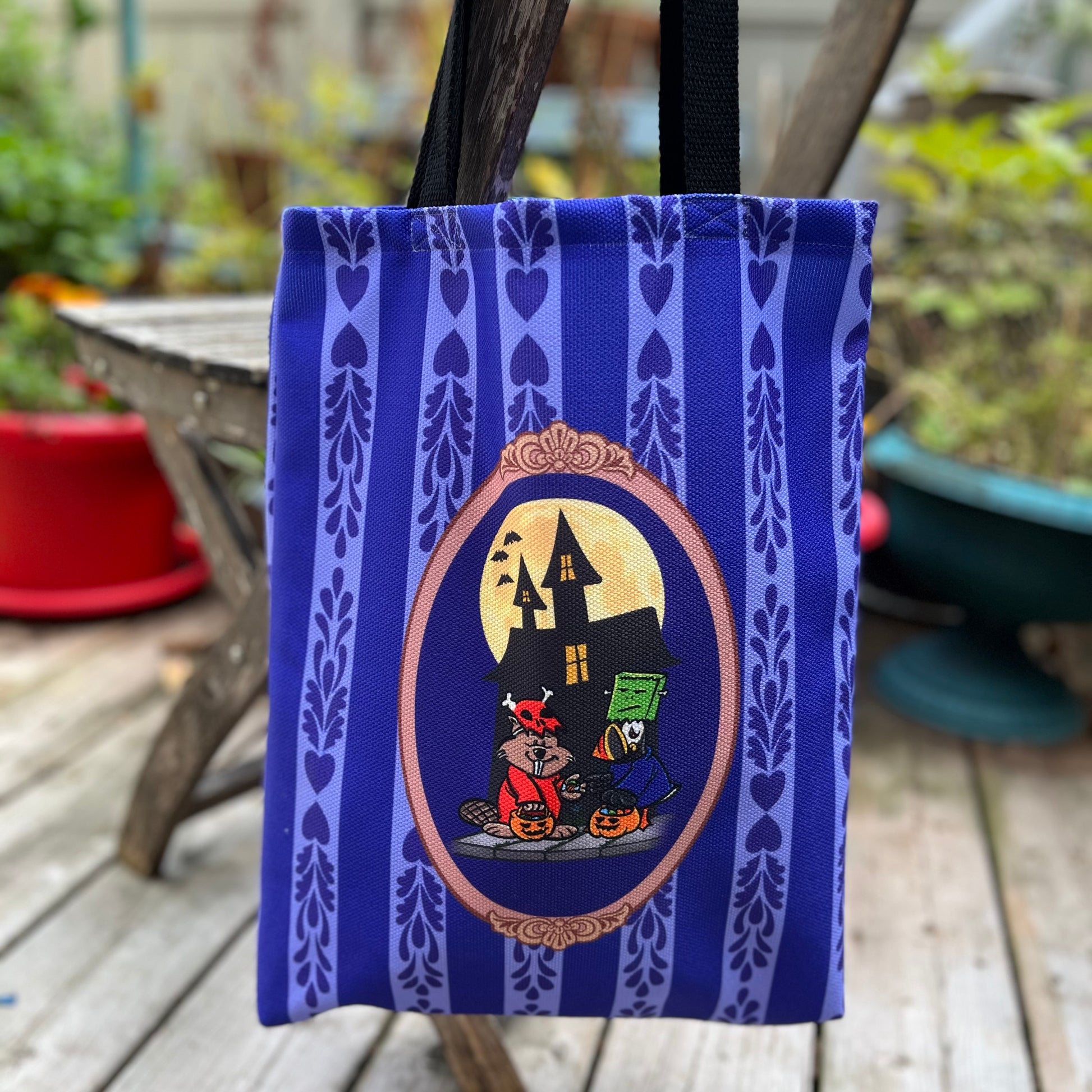 Blue tote bag with Halloween-themed design featuring a beaver and a puffin in costumes in front of a haunted house.