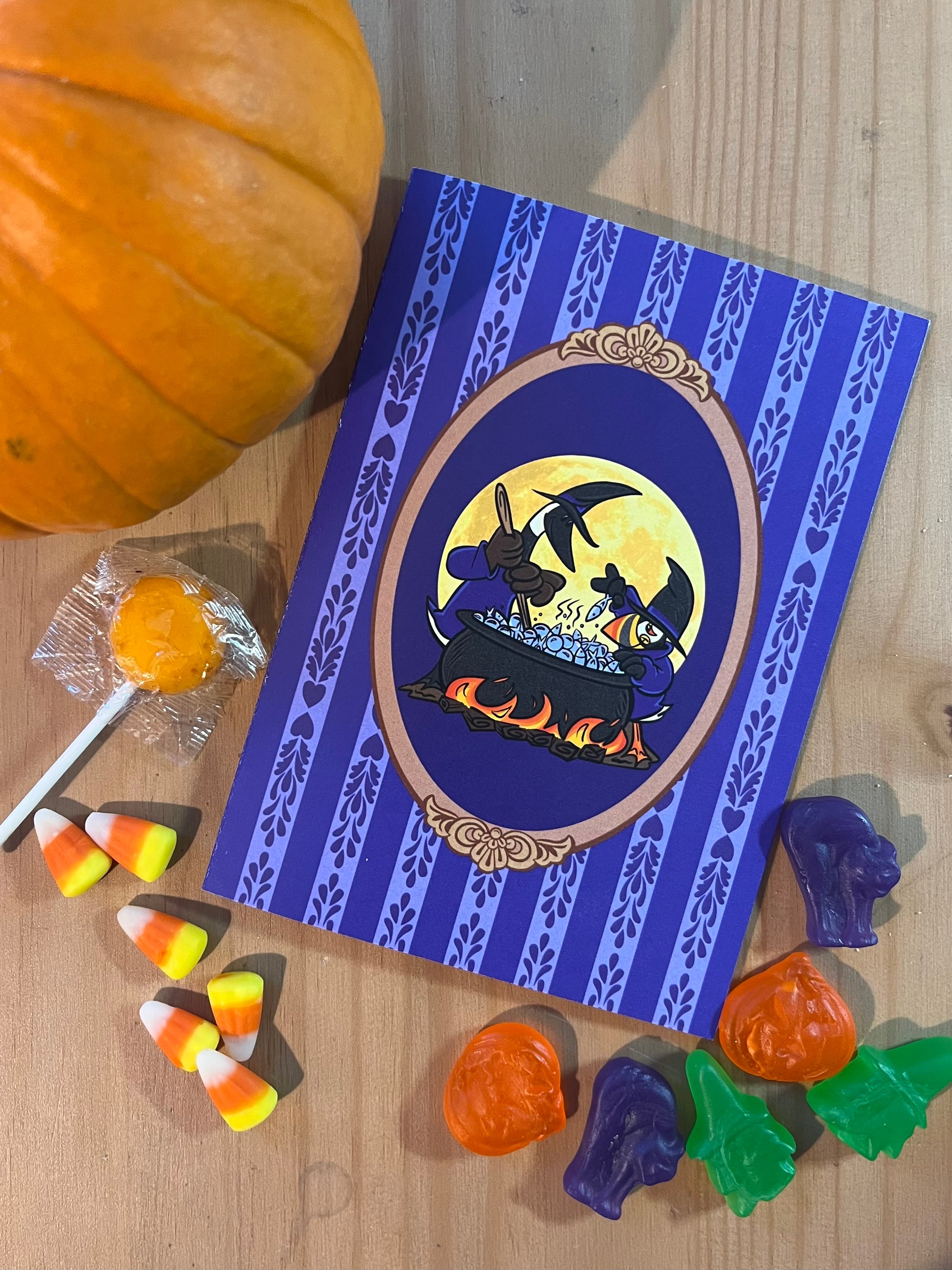 A notecard rests on a wooden surface, amongst a small pumpkin and Halloween candy. The notecard features two cartoon birds dressed as witches stirring a cauldron over a fire.