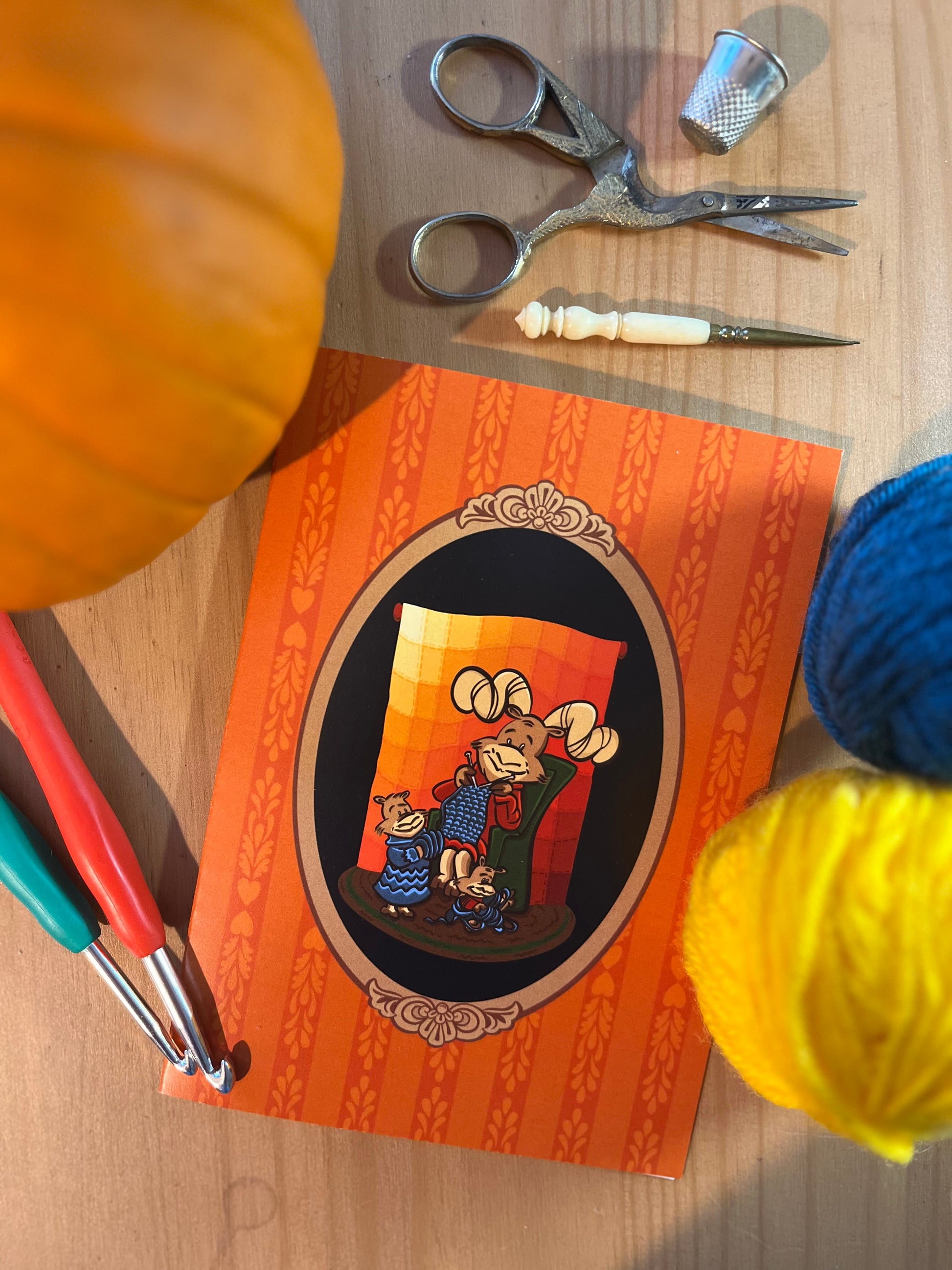 A notecard rests on a wooden surface surrounded by a small pumpkin and assorted sewing and crafting notions. The notecard features a cartoon big horn sheep in an armchair knitting in front of a quilt.