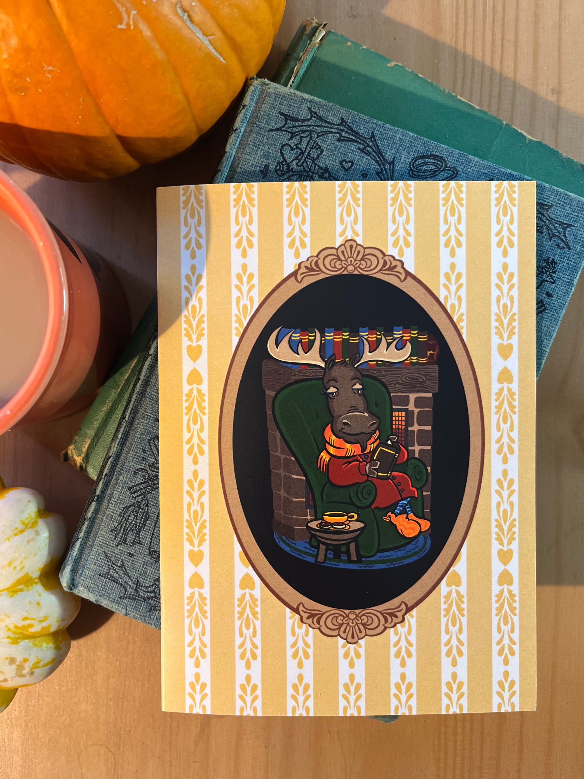 A notecard rests on a wooden surface, surrounded by a small pumpkin a cup of coffee and a stack of old books. The notecard features a cartoon moose reading in an armchair.