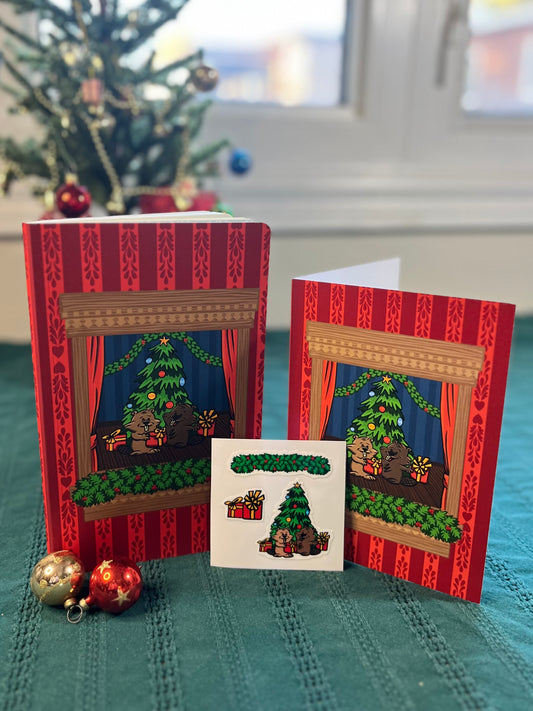 A greeting card, notebook and vinyl sticker sheet stand on display with a Christmas tree in the background. All three items feature a pair of cartoon beavers exchanging wrapped gifts.